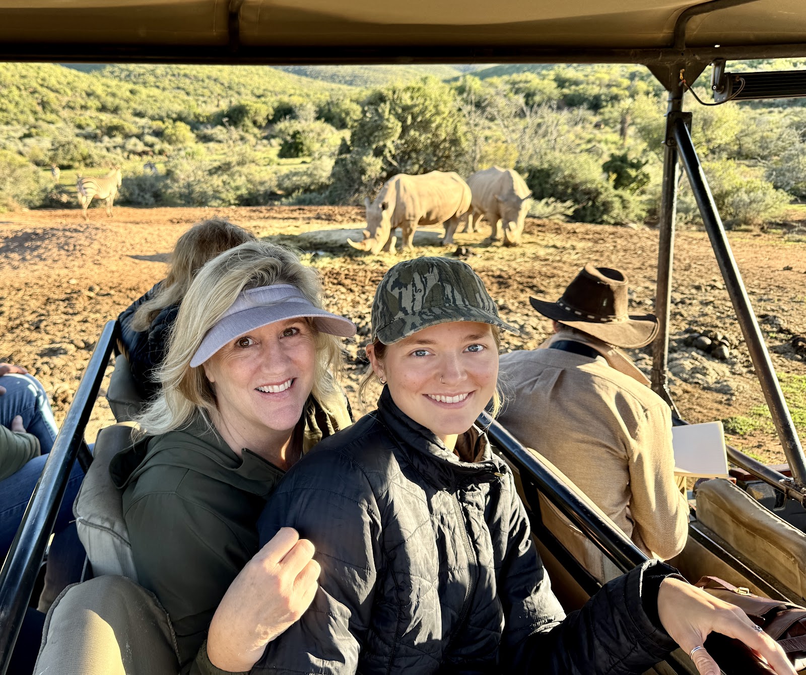 Two workshop travellers smiling in a safari vehicle with rhinos nearby